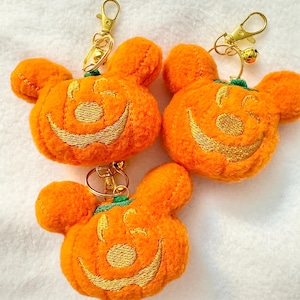 May include: Three orange pumpkin-shaped keychains with gold-colored hardware. Each keychain features a smiling jack-o'-lantern face embroidered in gold thread. The keychains have a Mickey Mouse-inspired design, perfect for Halloween.