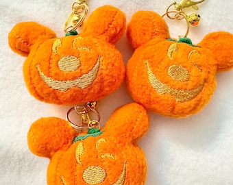Könnte beinhalten: Drei orangefarbene Kürbis-Schlüsselanhänger mit goldfarbener Hardware. Jeder Schlüsselanhänger zeigt ein lächelndes Jack-o'-Lantern-Gesicht, das mit goldfarbenem Faden bestickt ist. Die Schlüsselanhänger haben ein Mickey Mouse-inspiriertes Design, ideal für Halloween.