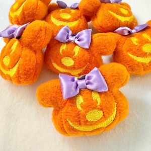 May include: A collection of orange plush pumpkin-shaped ornaments, each featuring a yellow embroidered jack-o'-lantern face and a small purple satin bow attached to the top. The plush items have rounded ears and a soft, textured fabric surface.
