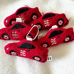 May include: A collection of red felt car-shaped keychains. Each keychain features black windows, white embroidered wheels, and the number "86" in white. The keychains have a white string for hanging.