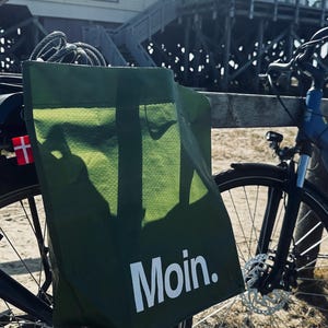 Könnte beinhalten: Eine dunkelgrüne, rechteckige Einkaufstasche mit dem in Weiß aufgedruckten Wort Moin., befestigt am Gepäckträger eines Fahrrads. Die Tasche hat eine gewebte Struktur und ein kleines rot-weißes Fähnchen an der Seite, platziert auf sandigem Boden.