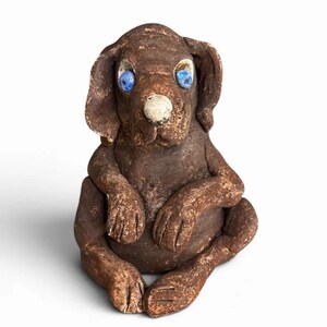 Könnte beinhalten: Eine braune Hundefigur aus Ton mit einer strukturierten, rustikalen Oberfläche. Die Skulptur hat zwei leuchtend blaue, kreisförmige Augen und eine markante weiße Nase. Der Hund sitzt in einer aufrechten Position mit den Vorderpfoten am Bauch.