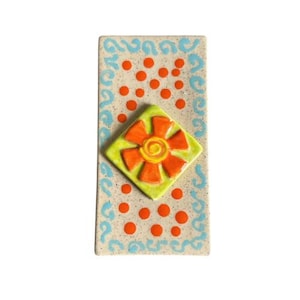 May include: A ceramic tile with a light brown background, blue and white swirls around the edges, and orange and yellow flower design in the center.