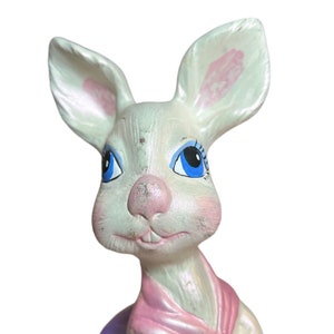 May include: A vintage ceramic bunny figurine with a white body, pink accents on the ears and nose, and blue eyes. The bunny has a pink scarf and a slightly distressed appearance, suggesting it is an older piece.