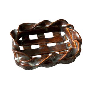 May include: A ceramic basket with a woven design. The basket is a dark brown colour with green highlights and a glossy finish. The edges are wavy, adding a unique touch. Ideal for holding small items. Measures approximately 20 cm in length.