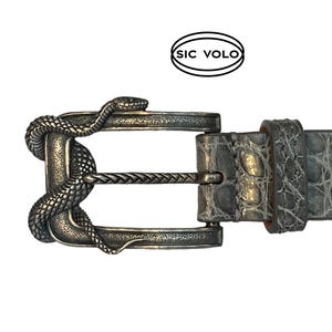 Puede incluir: Una hebilla de cinturón de plata con un diseño de serpiente. La hebilla está unida a un cinturón de piel de cocodrilo gris. El cinturón está etiquetado con "SIC VOLO".