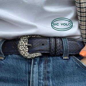 Puede incluir: Un cinturón de cuero azul oscuro con una hebilla floral plateada. La hebilla es un diseño grande y ornamentado con un patrón floral. El cinturón se lleva sobre un par de pantalones vaqueros azules. El texto "SIC VOLO" es visible en una camisa blanca.