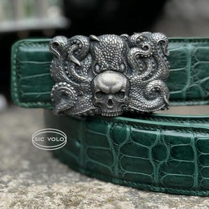 May include: A green alligator skin belt with a silver buckle featuring a detailed octopus and skull design. The buckle is engraved with the text "Sic Volo".