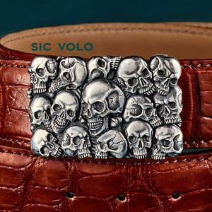 以下が含まれることがあります： 茶色の革ベルトに、銀色のスカルバックルが付いています。バックルには、詳細なスカルのクラスターがあります。ベルトは、クロコダイルのような質感のパターンです。「SIC VOLO」の文字が見えます。