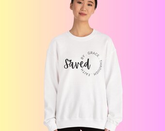 以下が含まれることがあります： 白いスウェットシャツ。筆記体で「Saved」の文字が書かれ、「By Grace Through Faith」が円状に配置されています。グラデーションの背景の前に、そのスウェットシャツを着た人がいます。