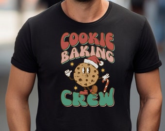 Pode incluir: Uma camiseta preta com um gráfico retrô de um biscoito usando um chapéu de Papai Noel e segurando um bastão de caramelo. O texto "Cookie Baking Crew" está impresso em vermelho, verde e branco.