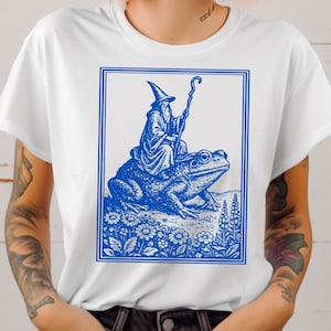 May include: White t-shirt featuring a blue illustration of a wizard riding a frog, set within a blue rectangular frame. The wizard wears a pointed hat and holds a staff. The frog is on a bed of flowers.