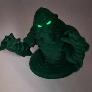 Puede incluir: Una figura verde impresa en 3D de un monstruo con ojos verdes brillantes. El monstruo está de pie sobre una base y tiene los brazos extendidos.