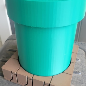 May include: A teal green pipe, similar to one from a video game, is placed on a brown brick-like base. The cylindrical pipe has a wide rim and a smooth surface. The base is made of individual rectangular blocks.