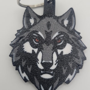 Puede incluir: Un llavero de cabeza de lobo negro y gris con ojos rojos. El pelaje del lobo es texturizado y el llavero tiene un anillo de metal plateado.