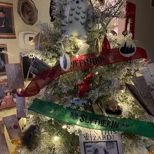 May include: A flocked Christmas tree decorated with Harry Potter themed ornaments, including a white owl, a red and gold ribbon with the words "Hufflepuff" and a green ribbon with the words "Slytherin". There is also a wanted poster with the text "Seen this wizard?" and a picture of a man with the text "Azkaban Prison 1391".