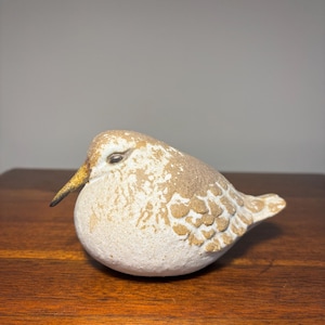 May include: A small, rounded bird figurine featuring a textured, off-white body with mottled tan and brown markings on the back and wings. The bird has a slender, downward-curving dark beak and a small, dark eye, with a smooth, plump shape.
