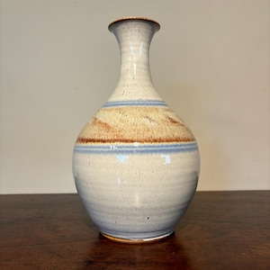 May include: A ceramic vase with a bulbous body and a slender neck. The vase features a light blue and tan color scheme with two horizontal blue stripes. The vase is approximately 10 inches tall.