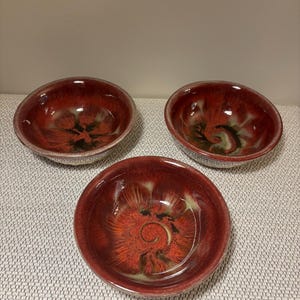May include: Three round ceramic bowls featuring a glossy glaze with a deep red and orange color palette. The interior surfaces display a swirling, radial pattern that transitions into darker tones toward the center, with a smooth, reflective finish.