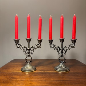 May include: Two matching metal candelabras with an antique brass finish, each featuring three arms and a fluted base. Each arm holds a slender, vibrant red candle. The metalwork displays intricate scrollwork and decorative detailing.
