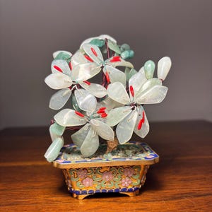 May include: A decorative bonsai tree sculpture featuring translucent pale green stone petals and bright red stamens. The tree is set in a rectangular metal planter with intricate blue, green, and pink floral enamel patterns and gold-toned trim.