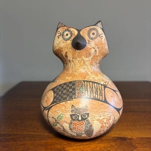 May include: A gourd vessel shaped like an owl with a marbled tan and brown surface. The body features black geometric patterns and a detailed illustration of a smaller owl perched on a branch with green leaves and orange accents.