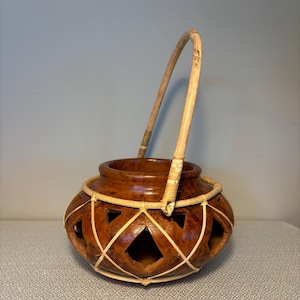 May include: A brown ceramic pot with a woven handle and intricate cutouts. The pot is decorated with a woven pattern of light brown strips.