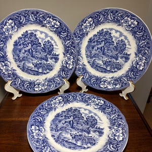 May include: Three round ceramic plates featuring a blue and white transferware pattern. The center of each plate depicts a rural landscape with a horse-drawn cart, while the border displays a detailed floral motif with leaves and berries.