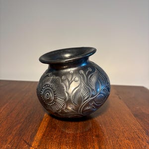 May include: A dark gray, handcrafted ceramic vase with intricate floral and leaf patterns. The vase has a rounded body and a flared rim. The detailed design is embossed on the surface, creating a textured appearance. The vase is displayed on a wooden surface.