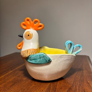 May include: A ceramic bowl shaped like a chicken with a white body, a bright orange comb and wattle, a yellow interior, and teal-colored wings and tail feathers. The chicken features a black beak and textured details on the neck and wings.
