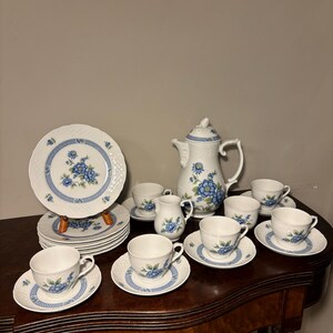 May include: A vintage porcelain tea set with a blue floral design. The set includes a teapot, cups, saucers, plates, and a creamer. The white porcelain is accented with blue floral patterns and a blue border. The set is arranged on a dark wooden table.