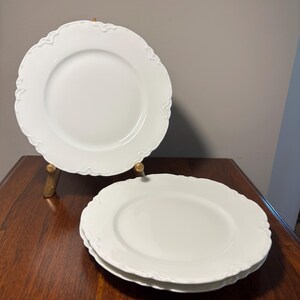 May include: A set of three white ceramic dinner plates featuring a smooth, glossy finish and a decorative, scalloped edge with subtle embossed detailing along the rim. One plate is displayed upright on a stand, while two are stacked flat.