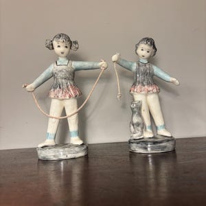 May include: Two ceramic figurines of children. One holds a jump rope, the other a cat. Both figures wear light blue tops, gray and red skirts, and white pants. The figurines stand on gray bases and are set on a dark brown surface.