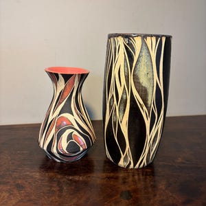 May include: Two decorative ceramic vases. The smaller vase has a red interior and a black, white, and red patterned exterior. The taller vase is black with a cream-colored, organic, linear design. Both vases are on a dark wooden surface.