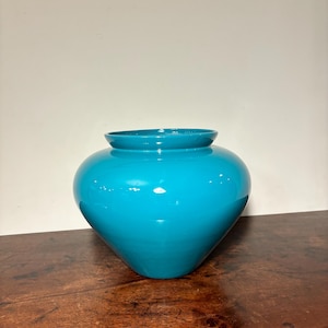 May include: A turquoise-colored vase with a glossy finish. The vase has a rounded body and a slightly flared opening. It is placed on a wooden surface against a plain white wall. The vase is a decorative item.