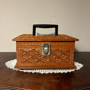 May include: A vintage wooden box with intricate floral carvings and a black handle. The box has a metal clasp and is set on a white lace doily. The box is brown and appears to be made of wood.