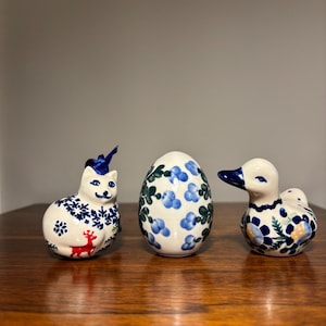 May include: Three ceramic figurines featuring a cat with a blue ribbon, a central egg, and a duck. Each piece is decorated with blue and green floral patterns, with the cat also displaying a small red deer and blue snowflake motifs.