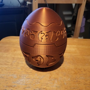 May include: A bronze-colored, egg-shaped object with intricate, carved patterns. The design features stylized symbols and decorative lines around the middle and top. The object is sitting on a wooden surface.