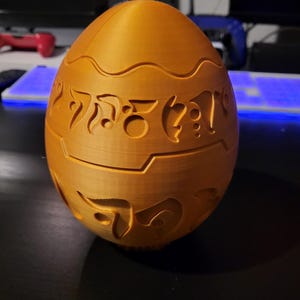 May include: A 3D-printed orange egg-shaped puzzle with intricate carved designs. The egg has a segmented design with decorative patterns and symbols. The surface has a textured appearance, and the egg is sitting on a dark surface.