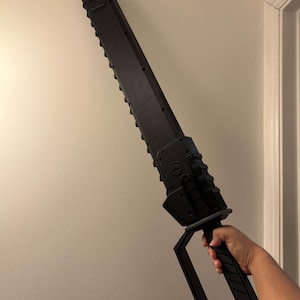 May include: A large, black prop sword with a serrated blade and a rectangular handle. The prop sword is held against a white wall. The handle has a rectangular opening for gripping.