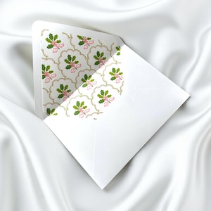 Könnte beinhalten: Ein weißer Umschlag mit einem gemusterten Innenraum. Das Muster zeigt grüne Stechpalmenblätter, rosa Schleifen und goldene Akzente auf weißem Hintergrund. Der Umschlag ist geöffnet und gibt den dekorativen Innenraum frei.