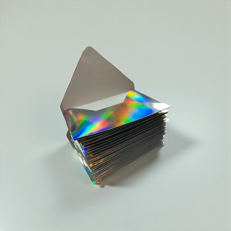 May include: A stack of iridescent, holographic paper cards, fanned out from a white envelope. The cards reflect a rainbow of colors, including green, blue, yellow, and purple. The envelope is open, revealing the cards.