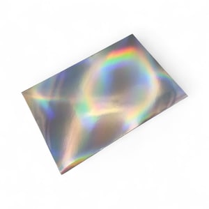 May include: A rectangular, iridescent package with a holographic finish. The surface reflects a spectrum of colors, including rainbow hues, against a white background. The packaging appears to be made of a shiny, reflective material.