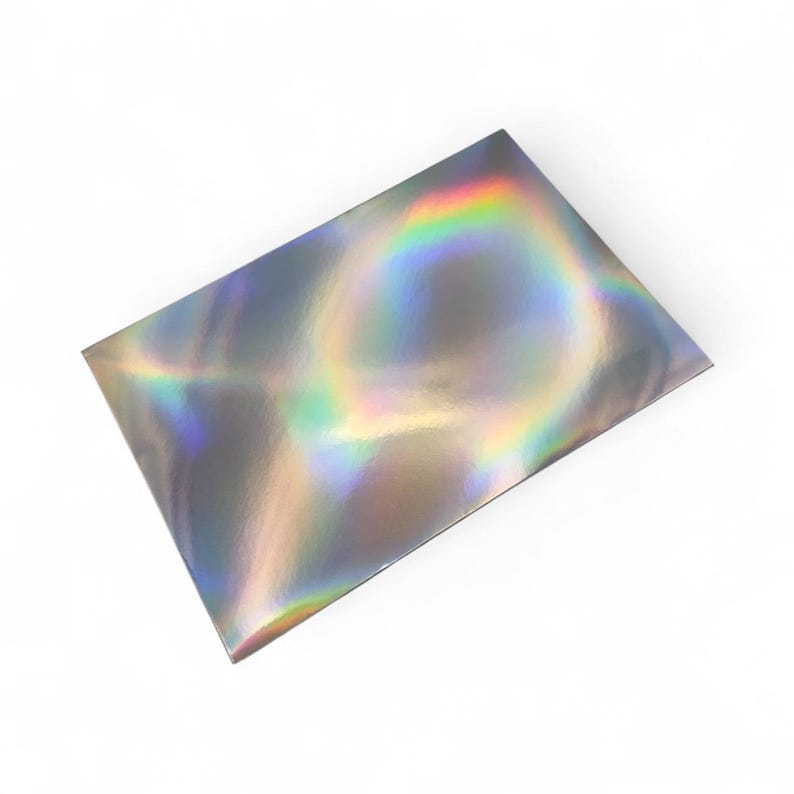 May include: A rectangular, iridescent package with a holographic finish. The surface reflects a spectrum of colors, including rainbow hues, against a white background. The packaging appears to be made of a shiny, reflective material.