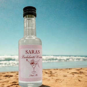 Op de afbeelding: Een heldere glazen fles met een zwarte dop, met het opschrift "Sara's Bachelorette Weekend" met een cocktailillustratie. De fles staat op een zandstrand met de oceaan en blauwe lucht op de achtergrond.