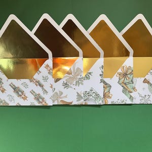 May include: A collection of decorative envelopes with gold foil interiors and white edges. The patterned exteriors feature festive illustrations of nutcrackers, gifts, and floral designs on a white background. The envelopes are arranged on a green surface.