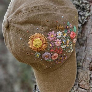 Puede incluir: Una gorra de béisbol de color tostado con intrincados diseños florales bordados a mano en el lateral. El bordado incluye un gran girasol amarillo, varias flores pequeñas rosas, blancas y moradas, hojas verdes y nudos franceses coloridos.