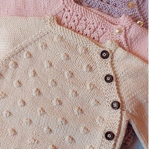 Könnte beinhalten: Drei Strickjacken für Babys in Pastellfarben. Die vorderste Strickjacke ist cremefarben mit einem strukturierten Muster und braunen Knöpfen. Die anderen beiden sind rosa und lavendelfarben, mit dekorativen Details und Perlen- oder Goldknöpfen.