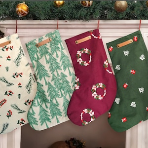 May include: Four Christmas stockings with festive patterns hang from a mantelpiece. The stockings feature designs of pine needles, Christmas trees, wreaths, and gifts. Each stocking has a leather loop for hanging and is in shades of green, burgundy, and cream.
