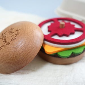 Peut inclure: Une boîte en bois avec les mots "Mom's Kitchen" gravés sur le couvercle. La boîte est ouverte et révèle un hamburger en feutre à l'intérieur. Le hamburger est fait de feutre rouge, jaune, vert et marron.
